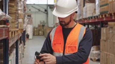 Güvenlik yeleği ve başlık takmış çalışkan bir erkek lojistik işçisi, büyük bir depodaki yüksek raflardaki envanterleri kontrol etmek için el tarayıcısı kullanıyor.