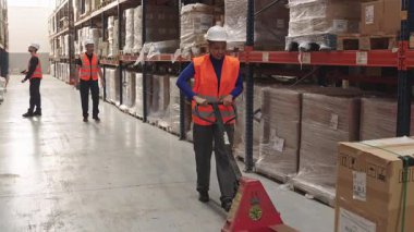 Geniş bir dağıtım merkezinde karton kutuları taşımak için manuel palet kriko kullanan güvenlik yeleği ve sert şapkalı genç bir kadın.