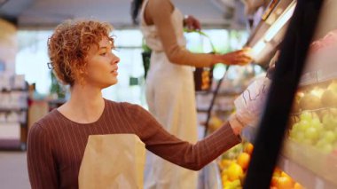 Kıvırcık saçlı, güzel bir kızıl kadın süpermarketteki raftan taze meyve topluyor. Bilinçli tüketici sağlıklı yiyecekler alıyor