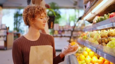 Kıvırcık saçlı genç bir kadın marketteki raftan taze portakal seçiyor. Marketten sağlıklı yiyecek almak isteyen müşteriler.