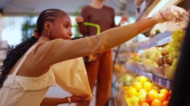 Young black woman carefully selecting a bunch of green grapes from a display shelf in a grocery stores fruit and vegetable department