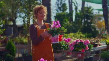 Tulumlu güzel kızıl saçlı kadın renkli saksı çiçeklerini dikkatlice seçiyor, onları güneşli bir bitkinin fidanlığında alışveriş arabasına koyuyor.