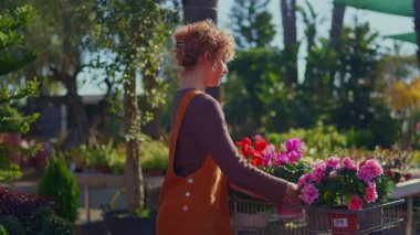 Kıvırcık saçlı genç bir kadın bahçe merkezindeki alışveriş arabasına saksı çiçeği koyuyor. Müşterilerin bitkileri seçerken yavaş çekim görüntüleri