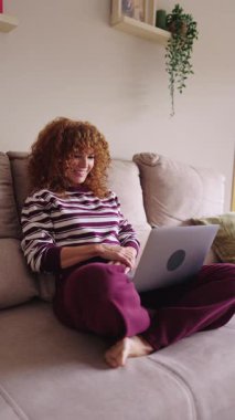 Kıvırcık saçlı genç bir kadın, oturma odasındaki video konferansında koltukta rahatça oturuyor, gülümsüyor ve bilgisayarına el sallıyor.