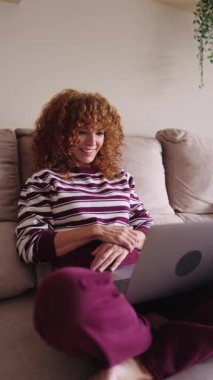 Kıvırcık saçlı mutlu kadın koltukta rahatça oturuyor, bilgisayarını video görüşmesi ve oturma odasından uzaktan kumanda ile arıyor.