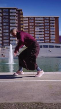 Tıraşlı saçlı, enerjik bir genç şehir parkında fıskiyeli ve arka planında binalar olan serbest dans figürleri yapıyor.