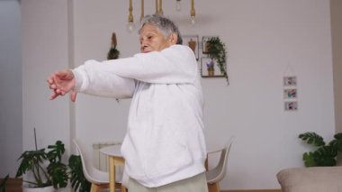 Sağlıklı, gri saçlı, oturma odasında egzersiz yapan yaşlı bir kadın. Aktif, olgun bir bayan sağlıklı ve zinde olmak için vücudunu esnetiyor.