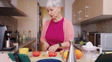 Kendinden emin son sınıf kadın yemek vlogcusu akıllı telefonuyla aşçılık dersi kaydederken sağlıklı bir yemek için taze malzemeler kesiyor.