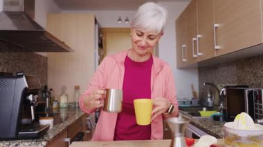 Aktif son sınıf öğrencisi, gri saçlı, mutfakta sütlü kahve hazırlayan bir kadın. Sabah rutini ve sağlıklı yaşam tarzı konsepti