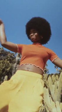 Doğal afro saçlı, neşeyle dans eden genç siyahi bir kadın. Renkli kıyafetler giyiyor ve açık mavi gökyüzünün altında gülümsüyor.