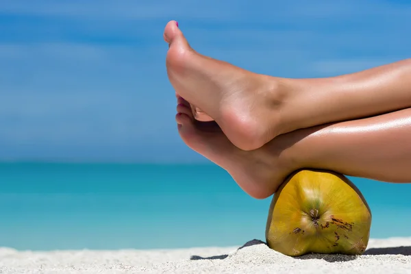 Female feet propped on coconut on the beach, blue sea background — Stock Photo, Image