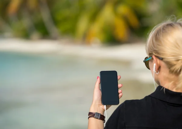 Mano de mujer mostrando un teléfono inteligente en blanco en la playa con el mar —  Fotos de Stock