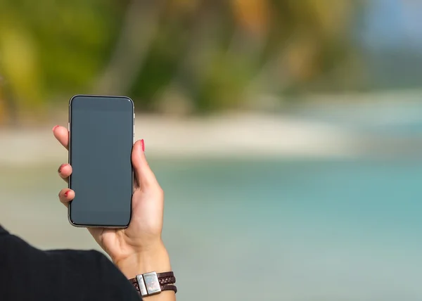 Mano de mujer mostrando un teléfono inteligente en blanco en la playa con el mar —  Fotos de Stock