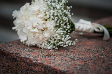 Beyaz çiçekli çiçek aranjmanı, bir kurdeleli düğün buketi ve bir taşın üzerinde duran iki altın yüzük. Düğün dekorasyonu, sanat eseri, çiçekçi.