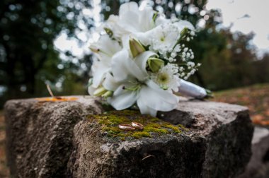 Beyaz çiçekli çiçek aranjmanı, bir kurdeleli düğün buketi ve bir taşın üzerinde duran iki altın yüzük. Düğün dekorasyonu, sanat eseri, çiçekçi.