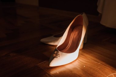 Gelinin beyaz ayakkabıları ve kahverengi arka planda bir çift altın alyans. Düğün aksesuarları