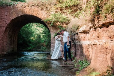 Yeni evliler, arka planda su ve taş kemer varken sarılıp öpüşürler.