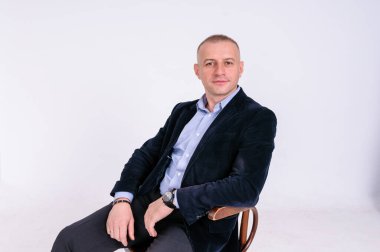 Mavi gömlekli, siyah pantolonlu ve mavi ceketli bir adam beyaz arka planda siyah bir taburede oturuyor.