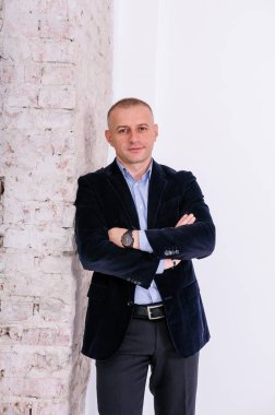 Mavi gömlekli, siyah pantolonlu ve mavi ceketli bir adam tuğla duvar arkasında duruyor.