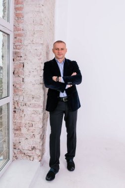 Mavi gömlekli, siyah pantolonlu ve mavi ceketli bir adam tuğla duvar arkasında duruyor.