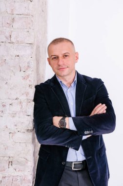 Mavi gömlekli, siyah pantolonlu ve mavi ceketli bir adam tuğla duvar arkasında duruyor.