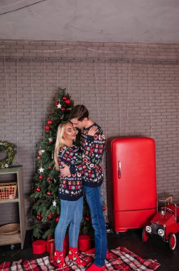 Elinde kazak ve kot pantolon giyen bir adam Noel ağacının arka planında sarılıp öpüşüyor ve kırmızı hediyeler veriyor.