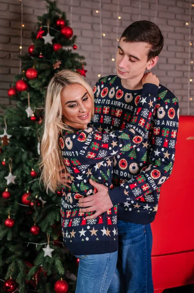 Парень с девушкой в свитере и джинсах обнимается на фоне елки и красных подарков — стоковое фото