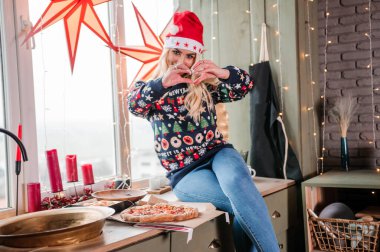 Kazaklı, kot pantolonlu ve Noel Baba şapkalı bir kız elinde bir dilim pizzayla masada oturuyor.