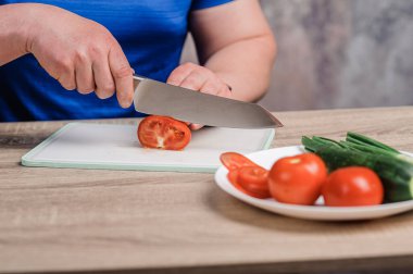A man cuts a red tomato on a board. Cucumbers and tomatoes in a white plate