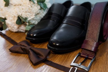 Elegant groom's essentials captured in a still life, showcasing polished brown leather loafers, a matching bow tie and belt beside a delicate white bouquet
