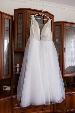 Hassas dantel korseli ve akıcı tüllü etekli güzel beyaz bir gelinlik, koyu renk ahşap bir dolabın önündeki askıdan nazikçe sarkıtılır ve etkinlikler için romantik bir düğün sahnesi yaratır.