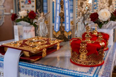 Yakın plan çekimlerde Ortodoks düğün ikonlarından alınmış dini eserler, mücevherli bir haç ve mavi kumaş kaplı bir masanın üzerine yerleştirilmiş kırmızı kadife bir taç sergilenir.