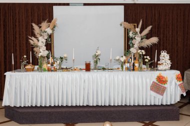 Güzel düzenlenmiş bir düğün masası, yemyeşil çiçekli süsler, narin otlar, altın mum tutacakları ve geleneksel Ukrayna işlemeli masa örtüsü.