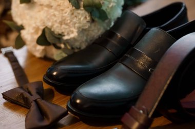 Close-up showcasing groom's black leather loafers, matching bowtie, and a white floral bouquet. luxury wedding accessories on wooden table, romantic mood, flat lay