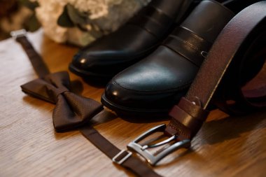 A close-up captures luxurious brown leather wedding shoes, a matching bowtie, and a belt neatly arranged on a polished wooden surface, evoking classic wedding style