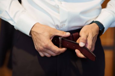 Koyu kahverengi bir kemeri donanma pantolonuna gümüş toka takarak ayarlayan bir adamın yakın plan görüntüsü resmi kıyafet ve düğün hazırlıklarını gösteriyor.