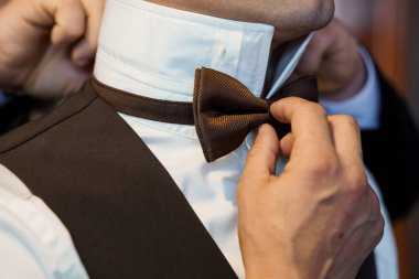 Yakın çekim, düğün hazırlıkları sırasında beyaz gömlek ve siyah yelek giyen bir damadın kahverengi papyonunu ayarlarken, zarafet ve inceliği yakalayan elleri resmediyor.