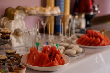 A lavish dessert table is beautifully arranged with a variety of treats, including colorful watermelon skewers adorned with green heart picks, delicate pastries, champagne glasses and elegant gold decorations, suggesting a festive celebration