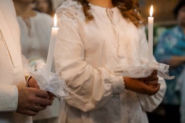 Ortodoks Hıristiyan nikahı sırasında mum tutan ellerin yakın çekimi, beyaz dantelli giysiler ve kutsal dini bir atmosfer sergileniyor.