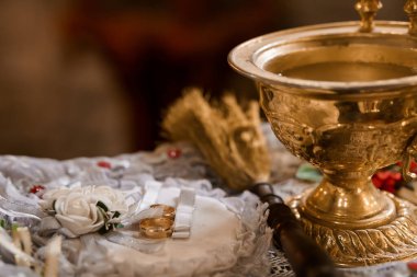 Intimate shot of an Orthodox Christian wedding ritual showing a golden chalice filled with water, wedding rings, delicate white roses, and ornate lace, symbolizing faith