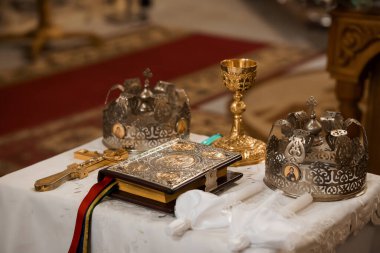 Ortodoks kilisesindeki resmi bir sahnede, taç giyme töreninde kullanılan altın taçlar, gümüş aksanlı kutsal bir kitap, altın bir kadeh ve haç gibi nesneler resmediliyor. Zengin kırmızı halı kutsallığı vurguluyor