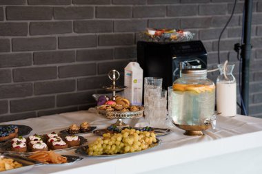 A beautifully arranged dessert table features tiered trays of pastries, fresh fruit, glasses of water, and a drink dispenser on a white tablecloth, set against a gray brick wall backdrop