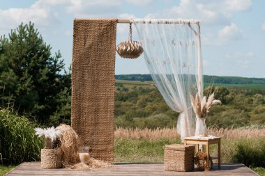 Güzel bir açık hava düğün kemeri jüt bir arka plan, dalgalı beyaz perdeler, pampa çim aksanları, ve güneşli bir arazide ayarlanmış ahşap mobilyalar.