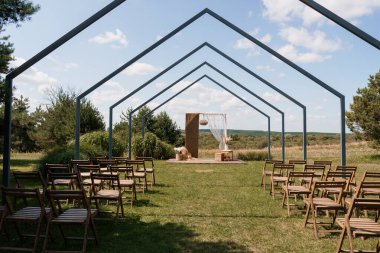 Çok güzel hazırlanmış bir açık hava düğün sahnesinde misafirler için ahşap sandalyelerle dolu sunağa uzanan bir dizi rustik ahşap kemer bulunur. dingin yeşil alan ve açık mavi gökyüzü barışçıl, doğal bir ortam yaratır.