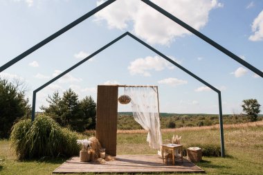 Güzel bir dış mekan düğün sahnesinde, çimenli bir tarlada, ahşap bir platforma kurulmuş, dokunmuş kumaşlarla süslenmiş üçgen bir metal kemer bulunur.