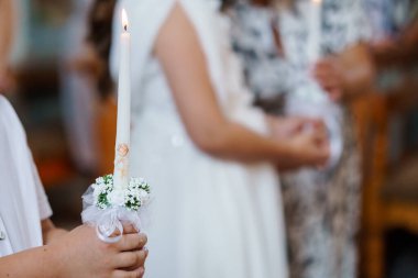 Beyaz çiçeklerle süslenmiş bir mumla süslenmiş bir oğlanı beyaz elbiseli bir kızın arka planına karşı göstererek dini geleneği ve inancı simgeleyen ilk komünyondan hassas bir sahne.