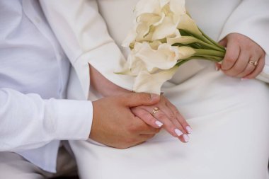 Evlilik teması için uygun, aşk, saflık ve evlilik bağlılığını sembolize eden bir buket beyaz zambak ve alyans tutan ellerin yakın çekimi.