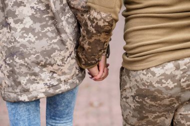 Kamuflaj ceketi ve pantolon giyerken el ele tutuşan iki kişinin yakın çekimi, desteği, ortaklığı ve ortak bir bağlantıyı sembolize ediyor, muhtemelen askeri personel veya sevgi dolu bir çifti.