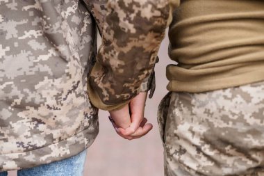 Yakın çekimde iki asker kamuflaj üniforması giyerken el ele tutuşurken görülüyor. Bu görüntü, askeri bir ortamda sevgi, destek ve yoldaşlığı temsil eden sessiz bir sevgi ve kişisel bağlantı hissi veriyor.