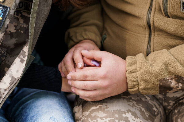 A close-up shot depicts two individuals clasping hands, conveying a sense of intimacy and support. one wears a camouflage jacket and the other denim, suggesting a personal connection with comforting tones